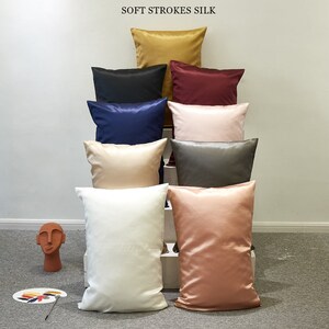 May include: A stack of eight pillowcases in various colors, including gold, black, navy blue, burgundy, pink, beige, gray, and peach. The pillowcases are made of soft silk and are arranged in a pyramid shape. The text "SOFT STROKES SILK" is displayed at the top of the image.