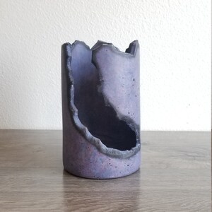 Könnte beinhalten: Eine zylindrische Vase aus lavendelfarbenem Beton mit einer gezackten, geodenartigen Öffnung. Der Innenraum ist dunkel, und die Ränder sind in einem dunkleren Farbton umrandet. Die Vase steht auf einer Holzoberfläche.