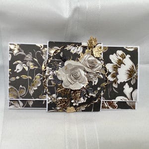 May include: A handmade greeting card with a floral design. The card features a dark gray background with gold and white floral patterns. A central embellishment includes paper roses and gold accents, creating a three-dimensional effect.