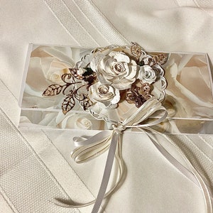 May include: A decorative white and gold floral clutch with a ribbon tie. The clutch features a floral design with paper roses and gold accents. The background of the clutch has a rose pattern.