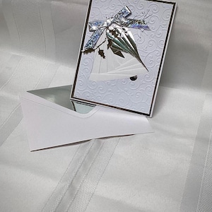 May include: A white greeting card with a silver bell design and a silver bow. The card has a textured background and a dark border. A matching white envelope with a silver interior is also present.
