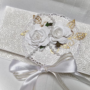 May include: A white, textured envelope with a floral design, adorned with white paper roses, gold-colored leaves, and a pearl strand. A white satin ribbon with gold trim is tied in a bow at the bottom, creating an elegant, handcrafted look.