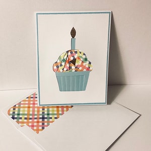 May include: A birthday greeting card featuring a cupcake illustration with a lit candle. The cupcake has a blue paper cup and a colorful, checkered frosting design. An envelope with a matching checkered pattern is also visible.