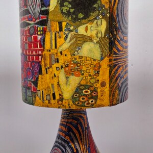 May include: A table lamp with a colorful, abstract design. The lampshade features a painting of two figures embracing, with a yellow, gold, and red color scheme. The base of the lamp is a swirling pattern of black, red, yellow, and blue.