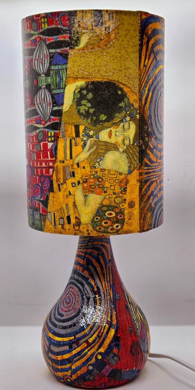 May include: A table lamp with a colorful, abstract design. The lampshade features a painting of two figures embracing, with a yellow, gold, and red color scheme. The base of the lamp is a swirling pattern of black, red, yellow, and blue.