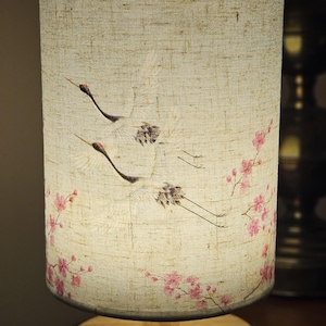 Könnte beinhalten: Zylindrische Tischlampe mit beigem Leinen-Lampenschirm. Der Schirm zeigt ein gemaltes Design von zwei fliegenden Kranichen und rosa Kirschblüten. Die Lampe hat einen runden Holzsockel und eine dunkelbraune Holzverkleidung.