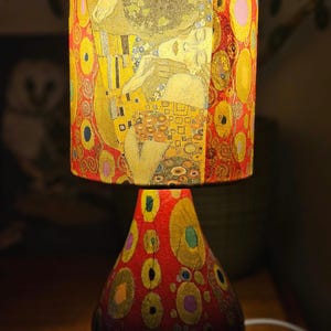 May include: A table lamp with a red and gold patterned base and a cylindrical lampshade featuring a painting of two figures embracing. The lampshade has a yellow, gold, and red colour scheme.