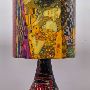 May include: A table lamp with a colorful, abstract base and a cylindrical lampshade featuring a painting of two figures embracing. The lampshade is decorated with a yellow, gold, and blue pattern.