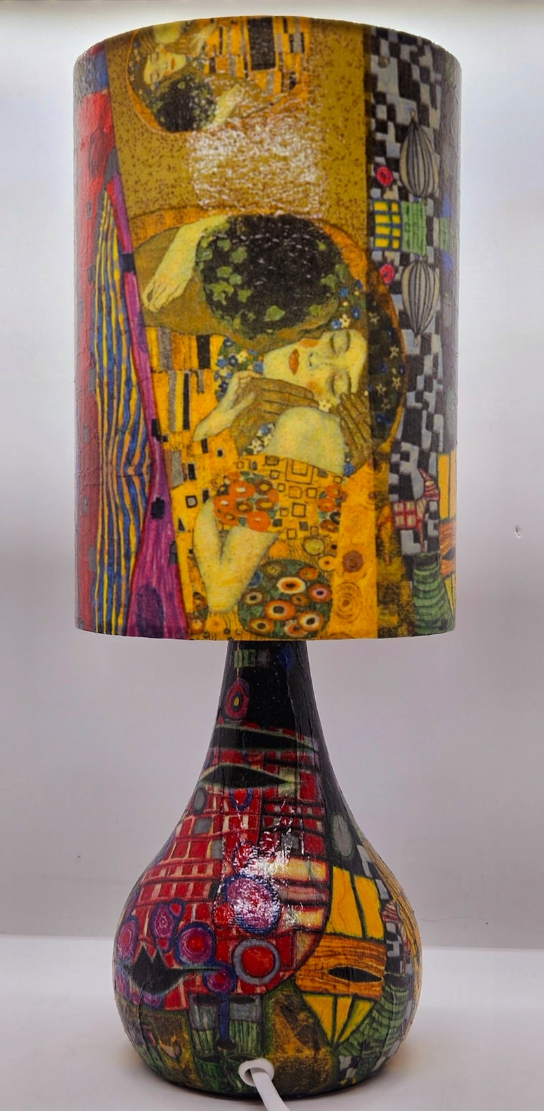 May include: A table lamp with a colorful, abstract base and a cylindrical lampshade featuring a painting of two figures embracing. The lampshade is decorated with a yellow, gold, and blue pattern.