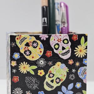 Puede incluir: Porta lápices cuadrado negro con un patrón colorido de calaveras de azúcar del Día de Muertos. El porta lápices tiene una variedad de flores, hojas y calaveras en tonos de rosa, amarillo, verde, azul y rojo.