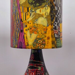 May include: A table lamp with a colorful, abstract design on the base and a cylindrical lampshade featuring a painting of two figures embracing. The lampshade is yellow, gold, and pink with a black and white geometric pattern on the side.