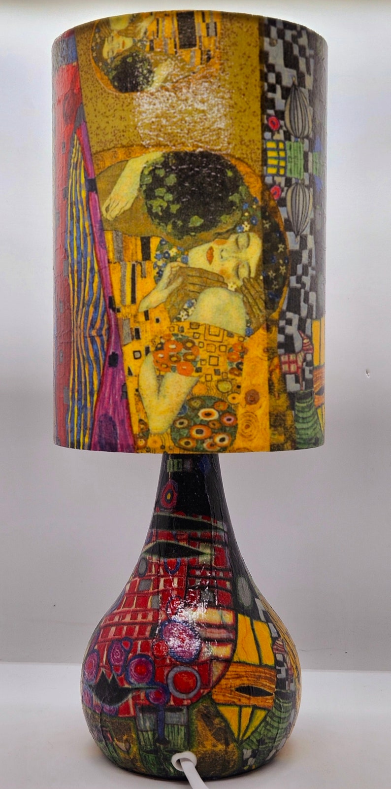 May include: A table lamp with a colorful, abstract design on the base and a cylindrical lampshade featuring a painting of two figures embracing. The lampshade is yellow, gold, and pink with a black and white geometric pattern on the side.