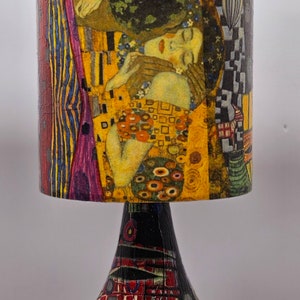 May include: A table lamp with a colorful, abstract design on the base and a cylindrical lampshade featuring a reproduction of Gustav Klimt's "The Kiss" painting.