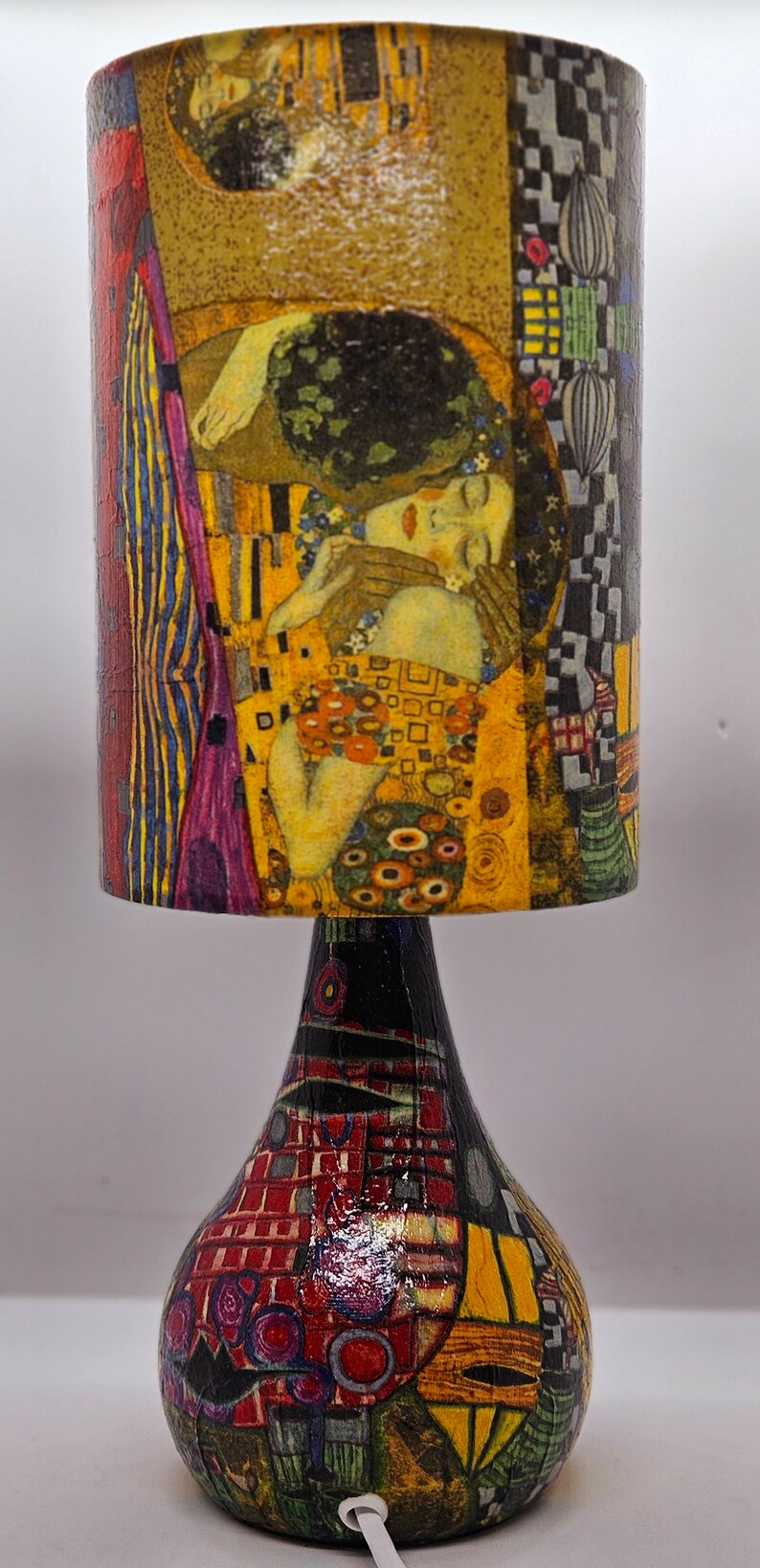 May include: A table lamp with a colorful, abstract design on the base and a cylindrical lampshade featuring a reproduction of Gustav Klimt's "The Kiss" painting.
