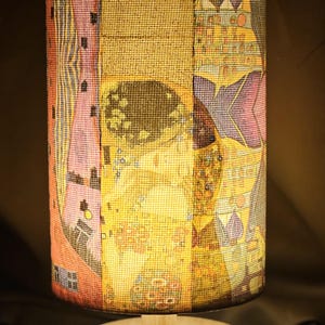 May include: A cylindrical table lamp with a patchwork lampshade. The shade features a collage of colourful, patterned fabrics in shades of yellow, pink, and purple. The lamp has a round wooden base and a wooden support.