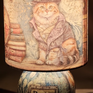 May include: A table lamp with a cylindrical shade featuring an illustration of a cat wearing a top hat and coat, alongside a stack of books. The base is decorated with a map pattern and a metal plaque with the text BECOME WHO YOU REALLY ARE.