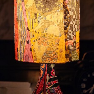 May include: A table lamp with a colorful, patterned base and a cylindrical lampshade featuring a reproduction of Gustav Klimt's "The Kiss" painting.