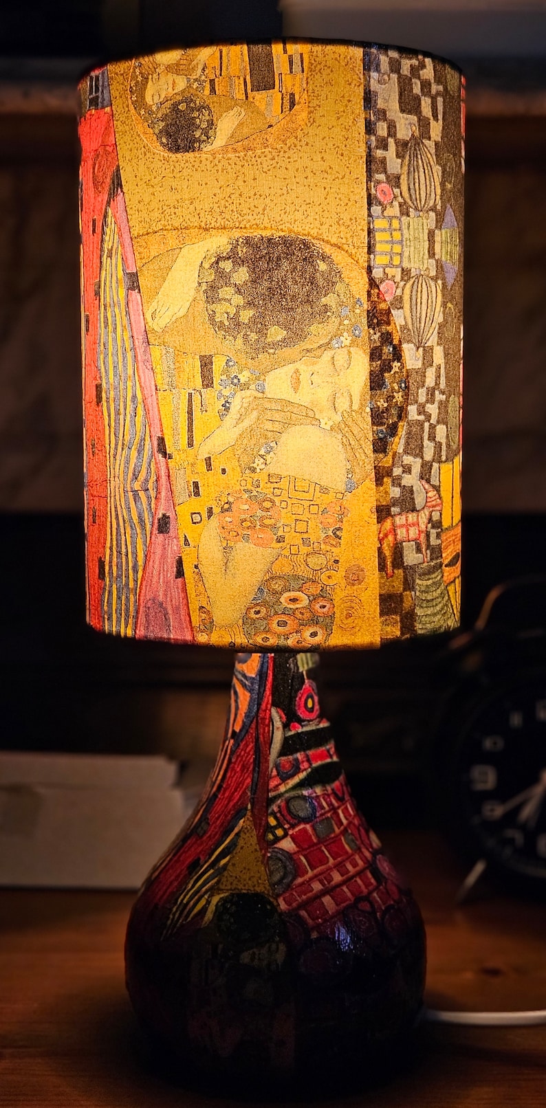 May include: A table lamp with a colorful, patterned base and a cylindrical lampshade featuring a reproduction of Gustav Klimt's "The Kiss" painting.