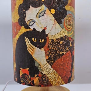 May include: A table lamp with a cylindrical linen shade featuring a colourful painting of a woman holding a black cat. The lamp has a wooden base.