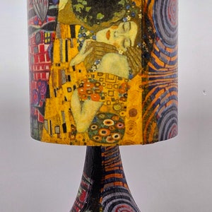 May include: A table lamp with a colorful, abstract patterned base and a cylindrical lampshade featuring a painting of two people embracing. The lampshade has a gold, yellow, red, and blue color scheme.