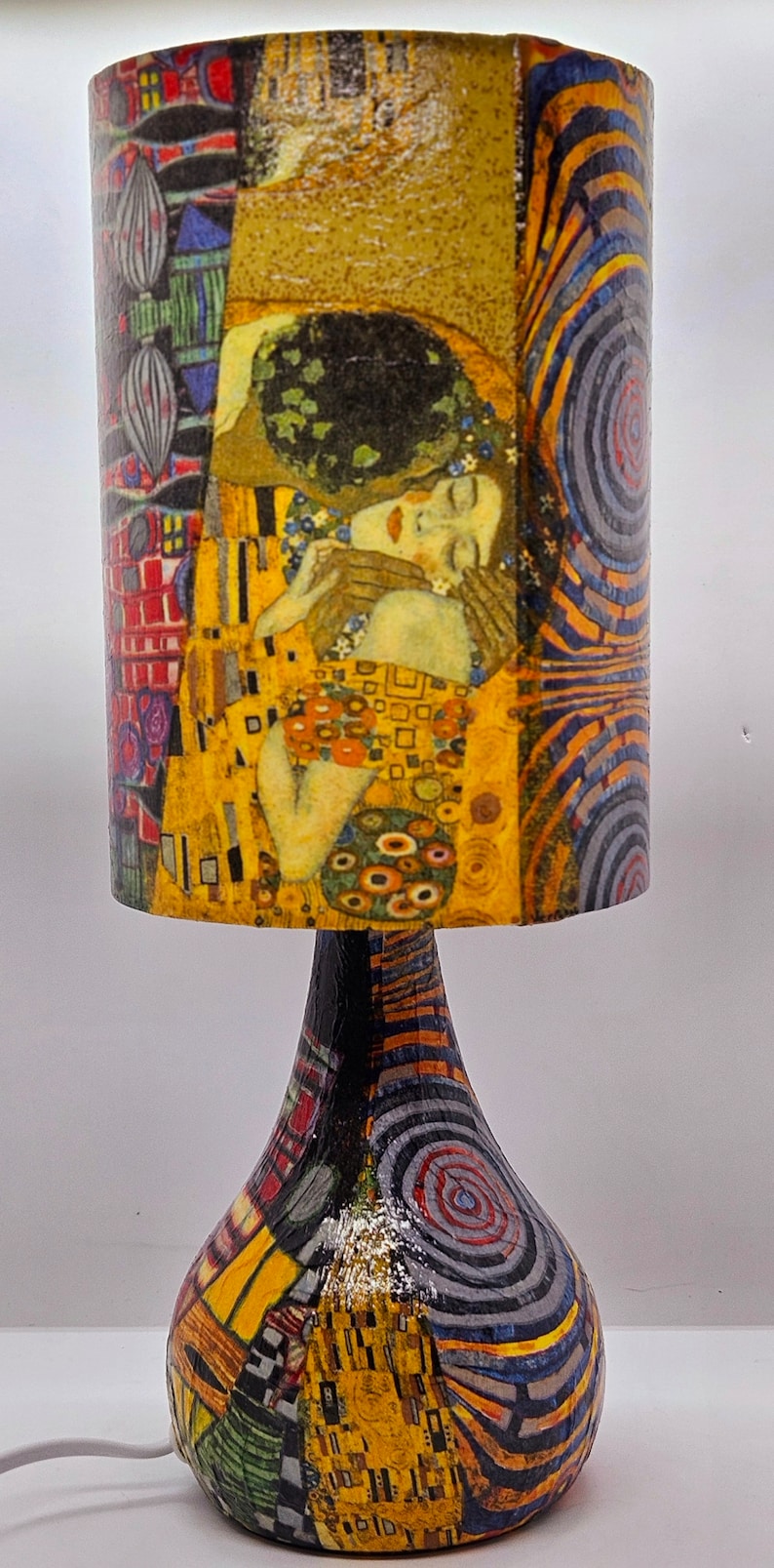 May include: A table lamp with a colorful, abstract patterned base and a cylindrical lampshade featuring a painting of two people embracing. The lampshade has a gold, yellow, red, and blue color scheme.