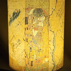 May include: A cylindrical table lamp featuring a printed design of Gustav Klimt's "The Kiss" artwork. The lampshade has a map-like background with the famous painting in the center. The base is made of light-colored wood.