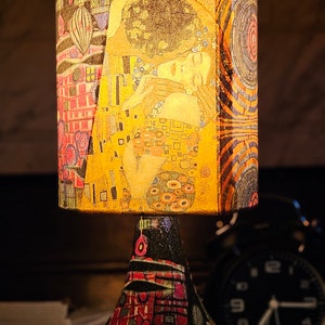 May include: A table lamp with a colorful, patterned lampshade featuring a painting of two people embracing. The lamp base is also decorated with a colorful, geometric pattern.