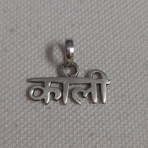 Pode incluir: Pingente de prata com a palavra "Kali" escrita em um script estilizado. O pingente tem uma argola na parte superior para uma corrente e é exibido em um fundo de tecido neutro.