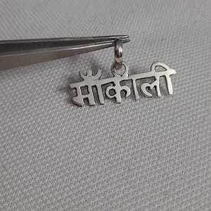 May include: A silver pendant with the Hindi word "माँ काली" (Maa Kali) engraved on it. The pendant is held by tweezers against a light gray textured background. The pendant has a small loop for a chain.