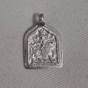 Può includere: Ciondolo in argento di forma rettangolare con un design a rilievo dettagliato. Il rilievo raffigura una figura che cavalca un leone, circondata da altre figure e motivi intricati. Il ciondolo ha un anello nella parte superiore.
