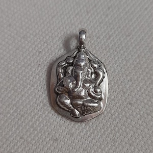 Può includere: Ciondolo in argento raffigurante la divinità induista Ganesha. Il ciondolo ha una forma esagonale con un anello nella parte superiore per una catena. L'immagine è su uno sfondo grigio chiaro strutturato. Gioielleria religiosa.