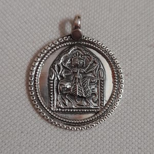 Può includere: Ciondolo color argento con un design in rilievo dettagliato. Il ciondolo rotondo presenta un'immagine centrale di una divinità o figura all'interno di una cornice ad arco, circondata da un bordo perlato. Un piccolo anello è in alto per una catena.