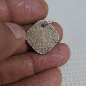 Może przedstawiać: Srebrzysty, kwadratowy wisiorek w kształcie monety z zaokrąglonymi rogami, zawierający napis INDIA, 1 ANNA oraz 1943. Na górnej krawędzi znajduje się małe metalowe kółeczko, dzięki któremu można go nosić jako zawieszkę przy naszyjniku lub bransoletce.