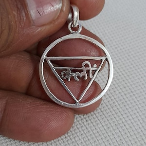 May include: A circular silver pendant featuring an inverted triangle frame enclosing the Devanagari script text "क्रीं". The pendant has a small silver bail at the top for attachment to a chain, showcasing a polished metallic finish throughout.