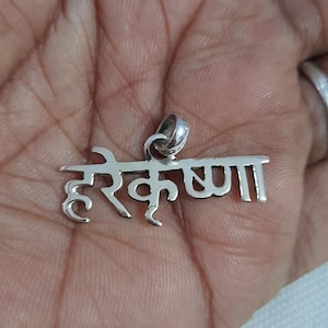 Peut inclure: Un pendentif en métal argenté arborant le texte en écriture devanagari "हरे कृष्ण", qui se traduit par Hare Krishna. Le pendentif présente une surface polie et réfléchissante, avec une petite bélière circulaire au sommet pour la chaîne.