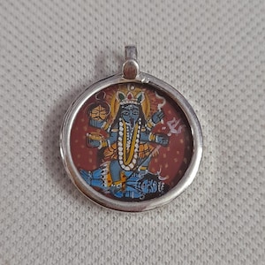 Könnte beinhalten: Ein runder silberner Anhänger mit einer handgemalten Miniaturillustration der hinduistischen Gottheit Kali. Die Figur ist mit blauer Haut, mehreren Armen und einem gelben Gewand dargestellt und steht über einer liegenden Figur vor rotem Grund.