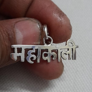 May include: A silver pendant with the Hindi text "महाकाली" (Mahakali). The pendant is in a stylized font and has a loop at the top for a chain. The pendant is held by a person.