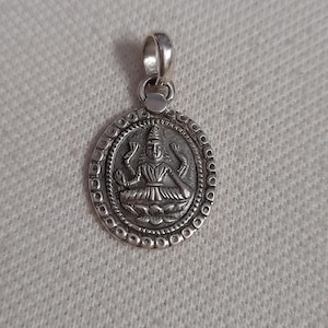 May include: A silver pendant featuring a detailed depiction of a seated deity within a circular frame. The pendant has a loop at the top for a chain and is set against a neutral background.
