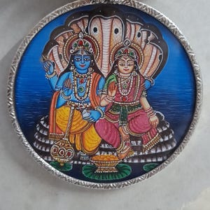 May include: Round pendant necklace featuring a detailed illustration of Hindu deities. The pendant has a silver-colored frame and a blue background. The necklace cord is made of purple and brown fabric with silver-colored beads.