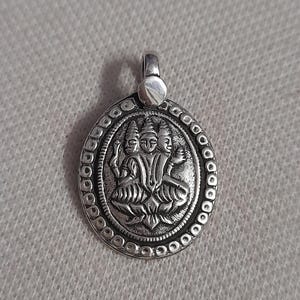 May include: Silver-toned pendant with a detailed relief of three figures within a circular frame. The pendant has a loop at the top for a chain and is set against a textured, light gray background. The design features intricate patterns.