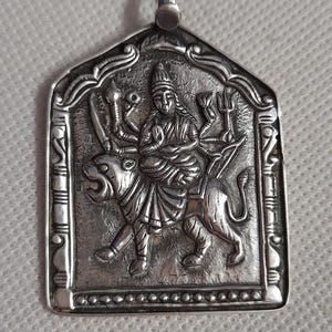Può includere: Ciondolo in argento raffigurante una divinità a cavallo di un leone. La figura ha più braccia e indossa un copricapo. Il ciondolo ha un bordo ornato e un anello per una catena.