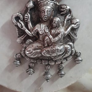 May include: Silver pendant necklace featuring a detailed depiction of a Hindu deity with multiple arms, sitting on a lotus throne. The pendant is adorned with intricate carvings and has small bells hanging from the bottom.