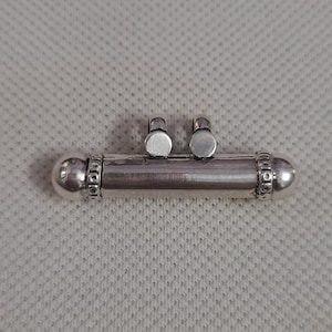 May include: A cylindrical silver pendant featuring a smooth central tube, rounded end caps with decorative beaded detailing, and two small circular loops positioned on the top edge for attachment to a chain or cord.