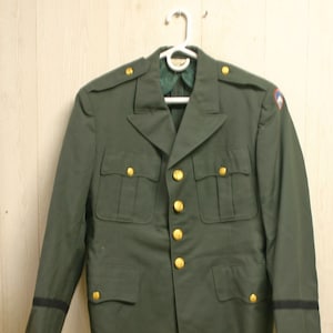 May include: A green military uniform jacket with gold buttons and a black stripe on the sleeves. The jacket has two pockets on the front and a patch on the right shoulder.