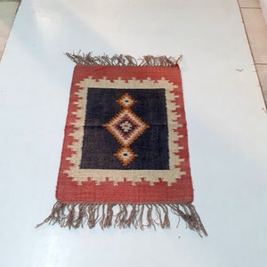 May include: A small rectangular rug with a geometric design. The rug features a central dark blue square with a diamond pattern in shades of orange, white, and red. The border is red with a white and red zigzag pattern. Brown fringe hangs from the bottom.