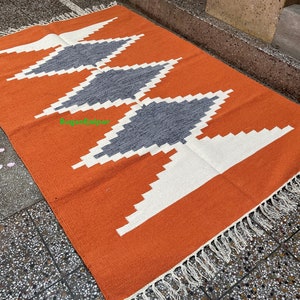 Puede incluir: Alfombra geom&eacute;trica naranja y blanca con formas de diamantes grises. La alfombra tiene un borde de flecos.