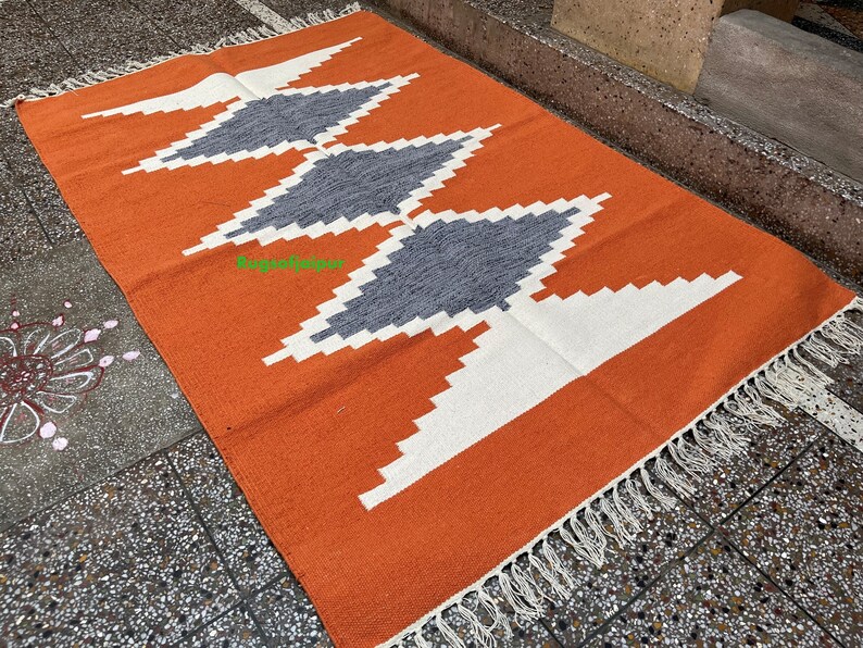 Puede incluir: Alfombra geom&eacute;trica naranja y blanca con formas de diamantes grises. La alfombra tiene un borde de flecos.