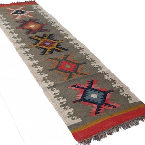 May include: A long, rectangular runner rug with a repeating geometric pattern. The rug features a central field of gray-green with colorful diamond-shaped motifs in red, yellow, blue, and orange. The edges are trimmed with a white zigzag pattern and red fringe.