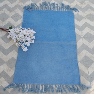 May include: A light blue cotton rug with fringe on all sides. The rug is laying on a white and gray tiled floor.
