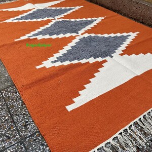 Puede incluir: Alfombra geom&eacute;trica con estampado naranja, blanco y gris con flecos blancos.